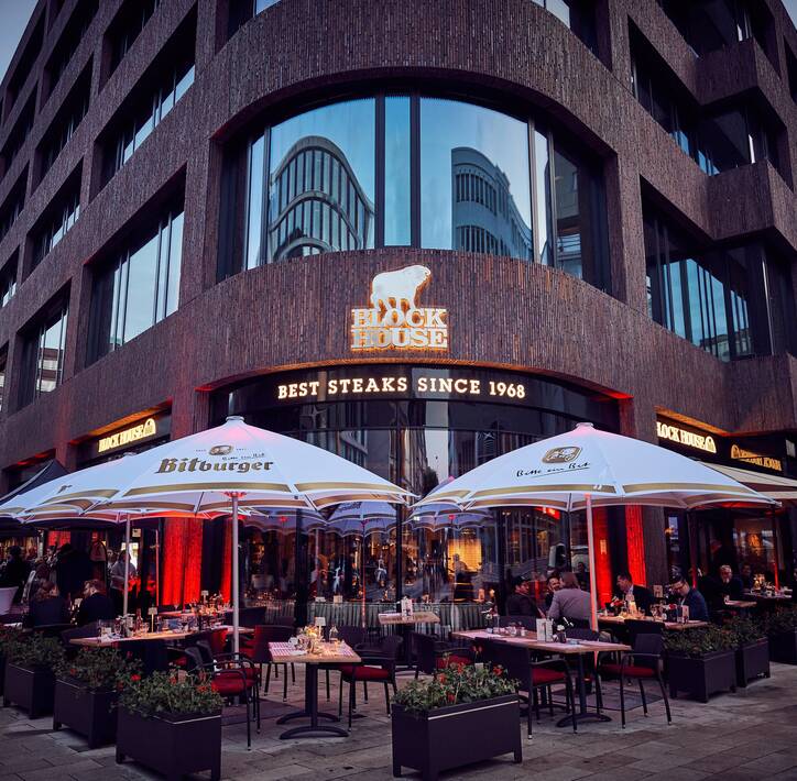 Außenansicht eines modernen Block House Restaurant mit einer Terrasse davor mit Tischen und Stühlen und Schirmen
