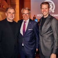 Drei Männer in formeller Kleidung stehen lächelnd zusammen bei einer Veranstaltung. Im Hintergrund ist eine Präsentation zu sehen.