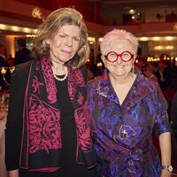 Zwei Frauen stehen in einem festlichen Restaurant. Die linke Frau trägt eine schwarze Bluse mit einem bunten Schal, die rechte Frau hat eine bunte Bluse und große rote Brille.