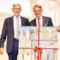 Zwei Männer stehen nebeneinander und halten ein gerahmtes Gemälde mit rotem Band. Der linke Mann trägt eine Brille und einen Anzug, der rechte Mann ist ebenfalls formell gekleidet.