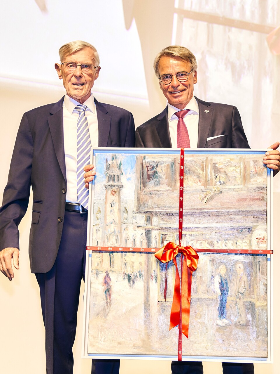 Zwei Männer stehen nebeneinander und halten ein gerahmtes Gemälde mit rotem Band. Der linke Mann trägt eine Brille und einen Anzug, der rechte Mann ist ebenfalls formell gekleidet.